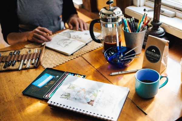Image description – A photo of a table with a mug, coffee plunger, pencils, paintbrushes and other artist tools, a person’s arms and hands are visible, and they hold a pencil in their hands with an open sketchbook in front of them