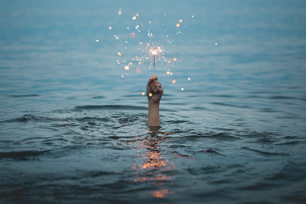 Image description – a hand reaches out from a large expanse of water holding a lit sparkler. The water is a grey/blue colour.