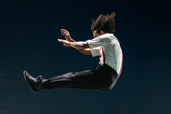 Image description – a photo of two performers. One is sitting on the  corner of a stage and the other is leaping high in the air above him.